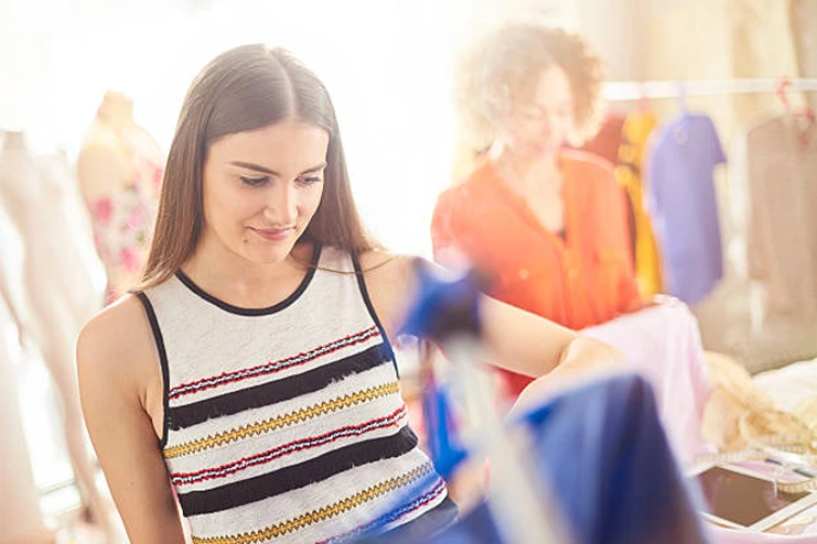 A woman looks at clothing in a brightly lit store, while another person in the background handles garments on a rack.