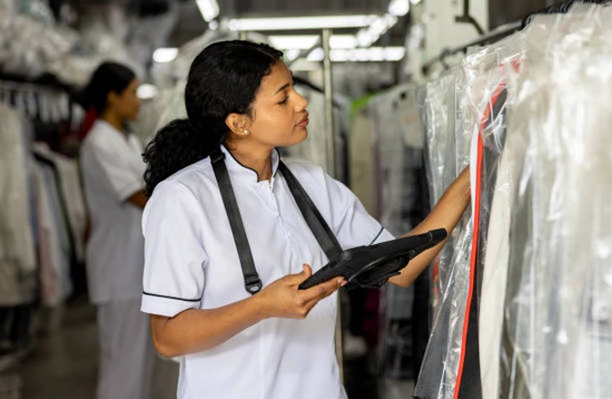 A woman in a uniform holds a tablet and inspects clothes covered in plastic at a laundry or dry cleaning facility. Another worker is visible in the background.