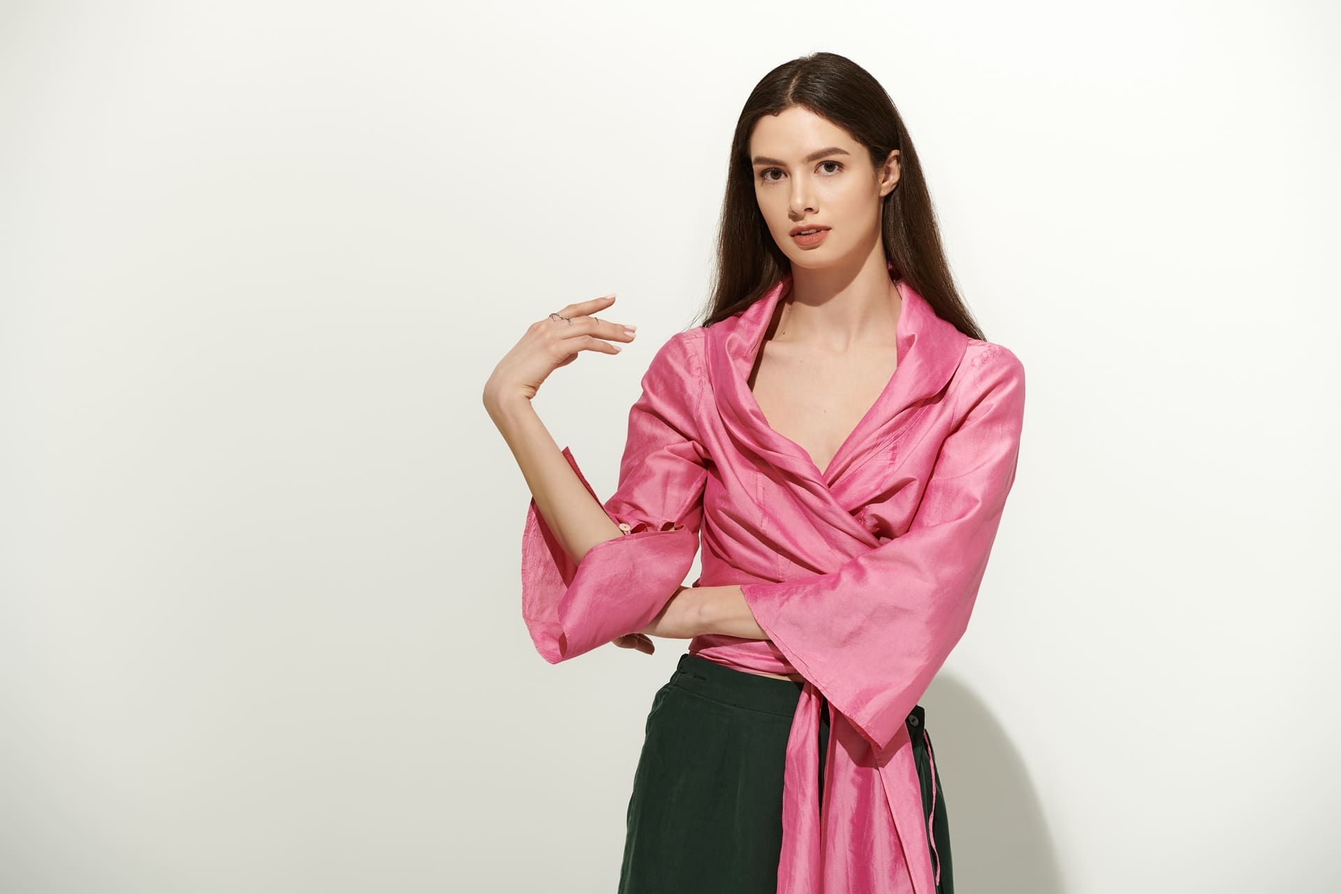 Woman with long brown hair wearing a shiny pink wrap top and dark green pants stands against a plain white background.