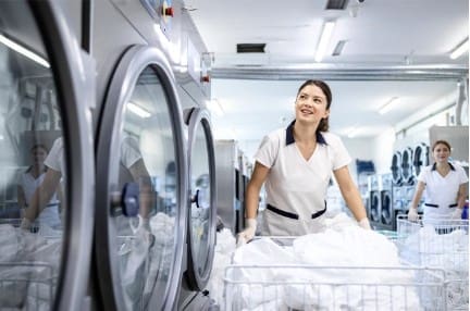 A woman in a laundry facility loads white linens into a washing machine, with other workers and laundry machines visible in the background.
